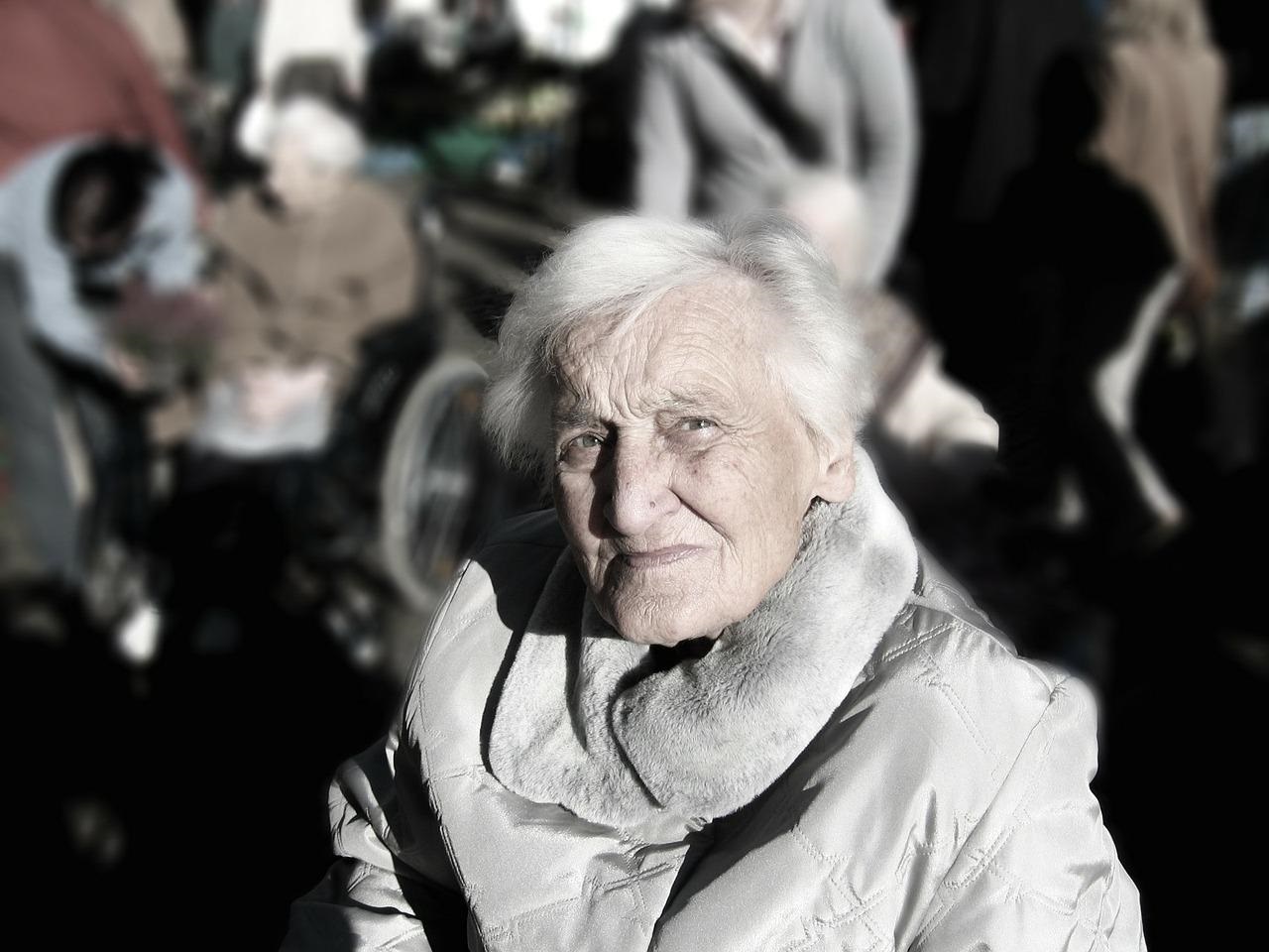 Ältere Dame mit kurzen, weißen Haaren blickt nachdenklich in die Kamera. Im Hintergrund sind weitere Personen zu erahnen.